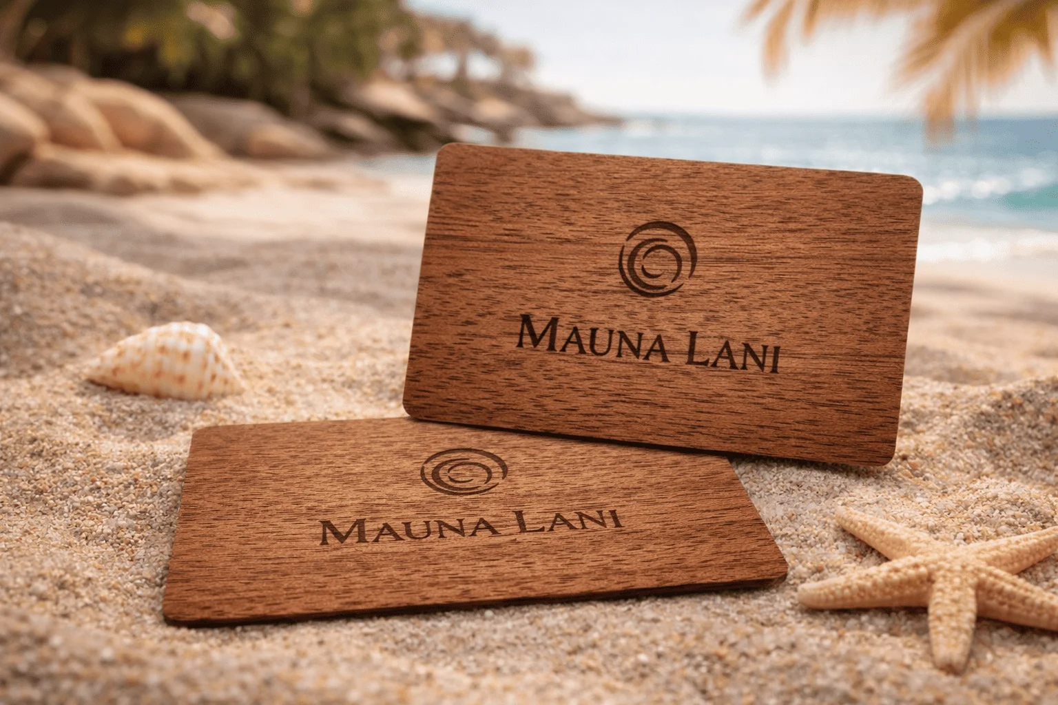 Wooden hotel key cards with "Mauna Lani" logo, resting on sandy beach with seashells and palm trees in the background.