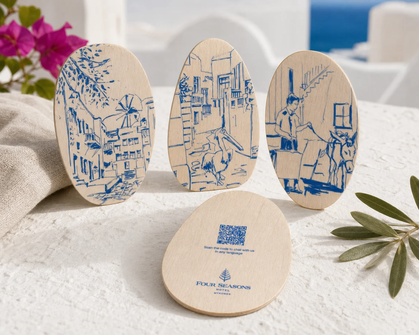 The four Four Seasons Mykonos pebble-shaped wooden key cards on a textured white table with bougainvillea and olive branch — windmill, pelican and donkey artwork faces plus the QR code back with the Four Seasons Hotel Mykonos logo