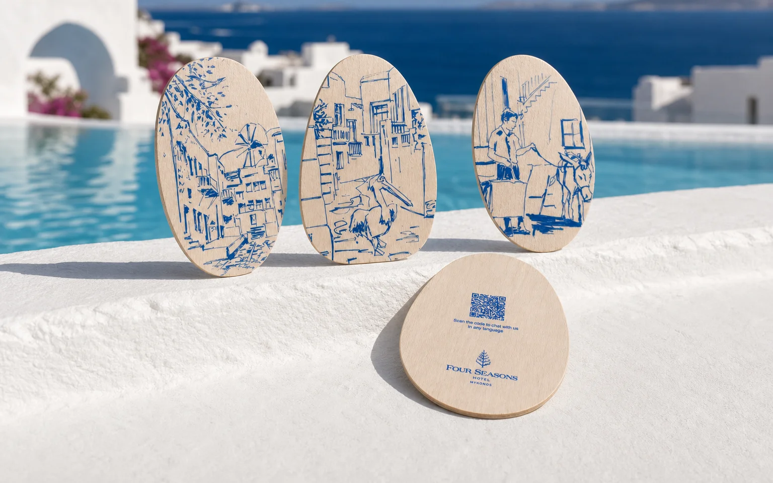 Four Seasons Mykonos pebble-shaped wooden key cards with blue island artwork standing on a white pool ledge above the Aegean Sea