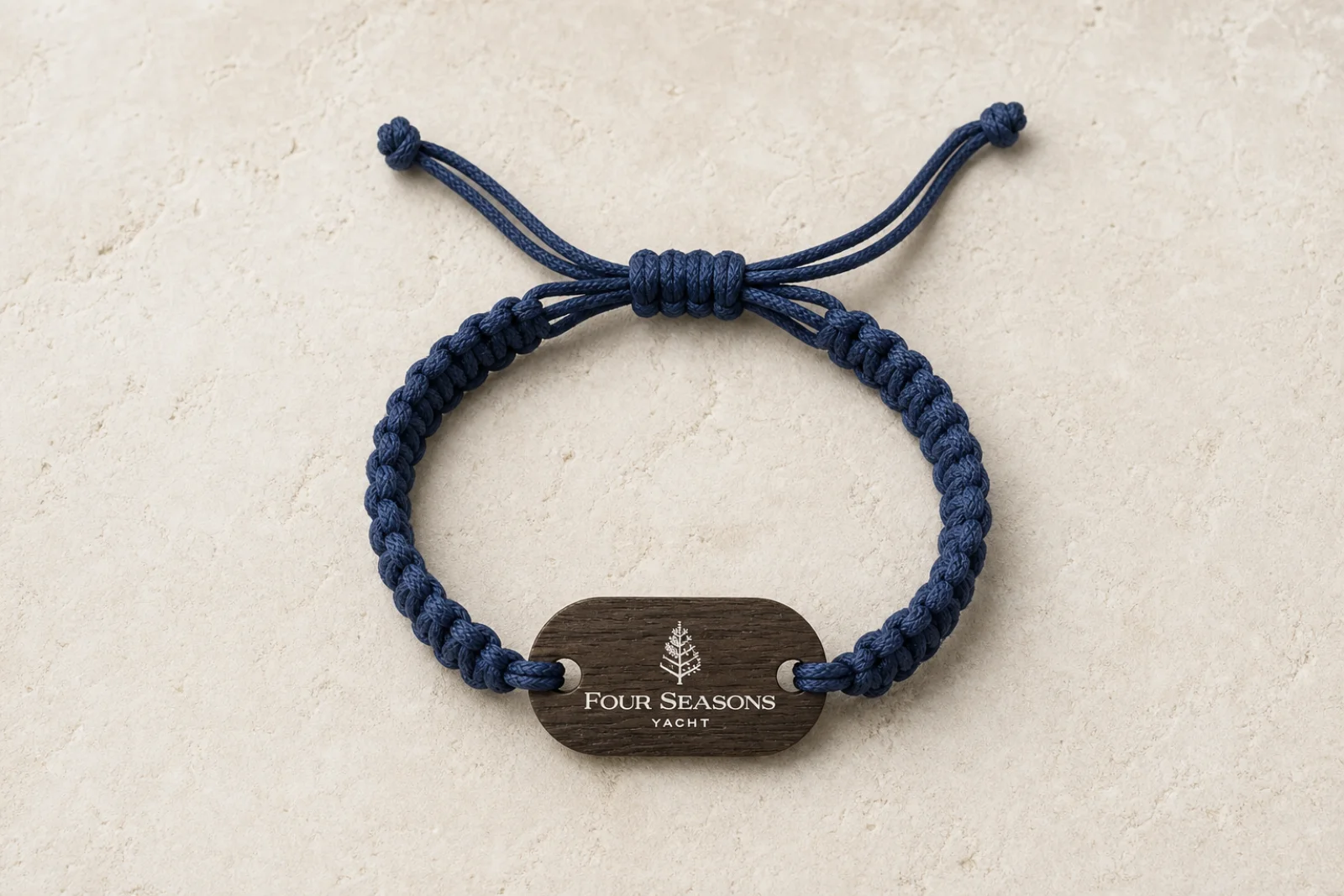 Flat lay of the Four Seasons Yacht wooden RFID wristband on a travertine stone surface, showing the full navy macramé weave and adjustable sliding knots