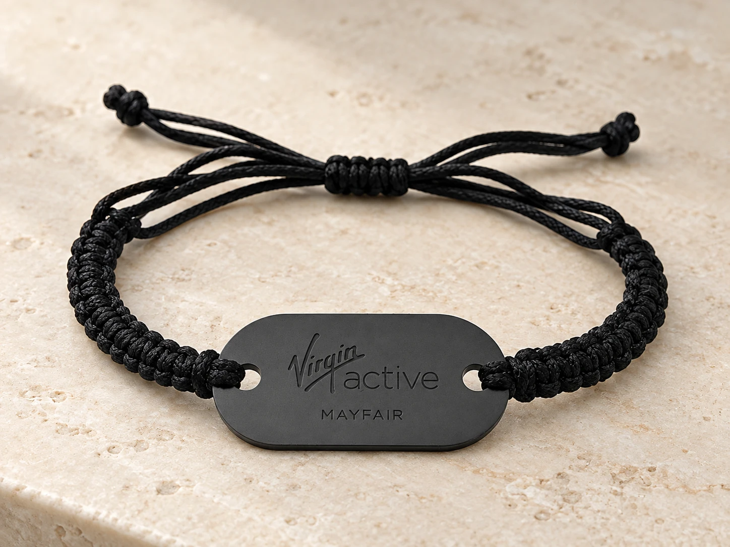 Virgin Active Mayfair custom wristband with a dark engraved tag and black braided cord on a stone counter